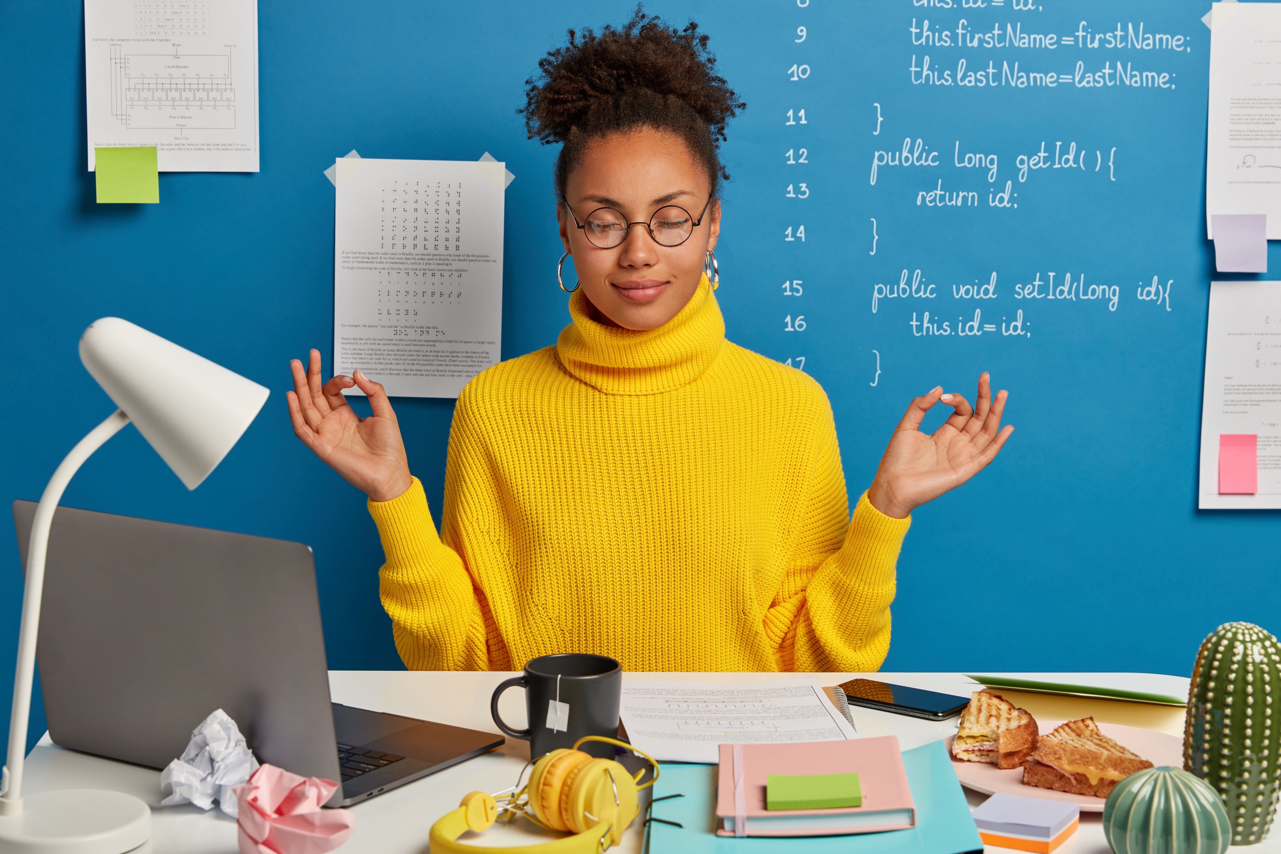 Young woman with glasses in a yellow sweater meditating in her job with a blue background